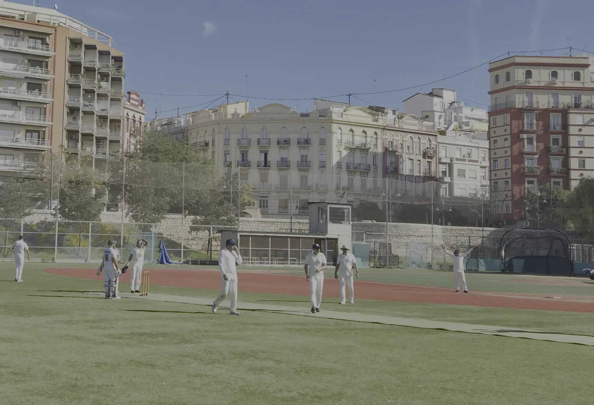 Cricketers in whites at the ground in the former Turia riverbed, with Valencia buildings behind